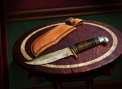 The Forgotten Age: A well-used and notched knife, about 6 inches in length, lies on an old wooden table. Its leather sheath lies next to it.