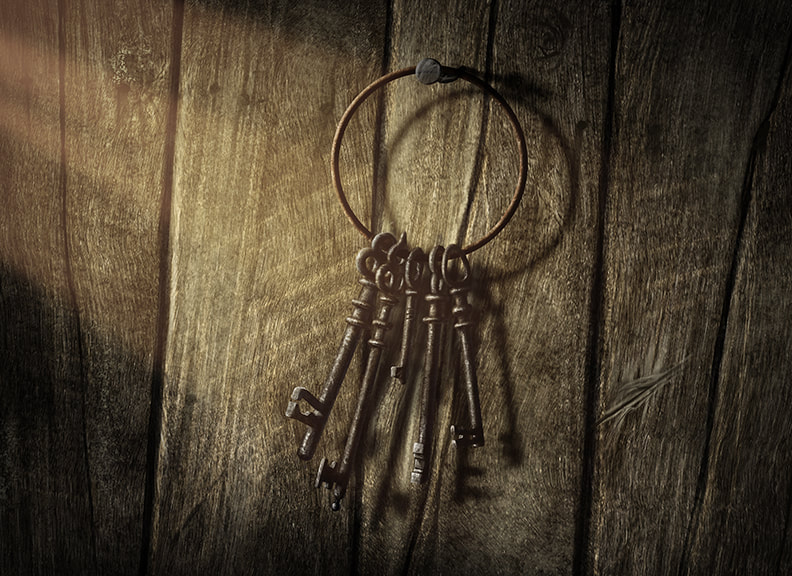 Several keys hang from a large, old brass keyring. The keyring hangs from a rusty hook on a wooden wall.